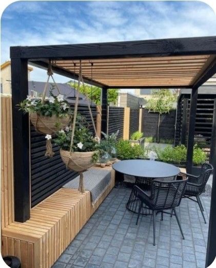 Pergola z czarnymi elementami konstrukcyjnymi i drewnianym zadaszeniem, wiszące donice z białymi kwiatami, drewniana ławka z szarą poduszką, okrągły stół i czarne krzesła na szarym brukowanym patio.