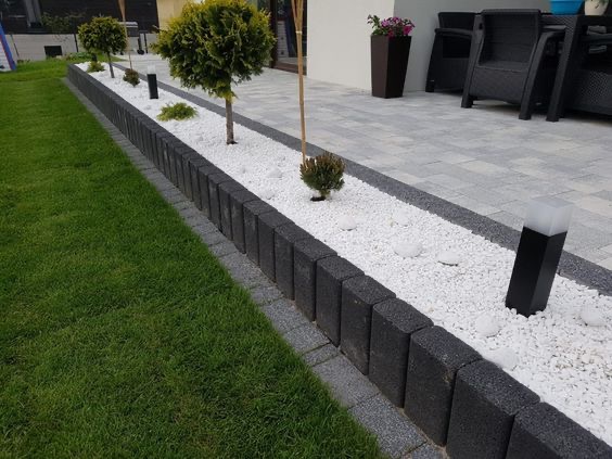 Ogród z niskim murkiem z ciemnej kostki, białym żwirem, małymi drzewkami i lampami wzdłuż ścieżki, trawnik po lewej stronie, szara kostka brukowa na tarasie w tle.