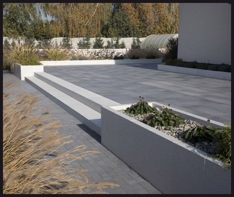 Nowoczesna przestrzeń zewnętrzna z szarej kostki brukowej, białych obrzeży i podwyższonych rabat z roślinnością ozdobną, w tle widoczne drzewa i fragment budynku.