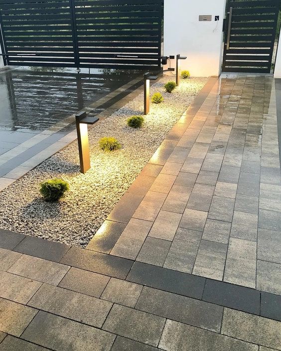 Ułożona kostka brukowa w odcieniach szarości, oświetlona lampami ogrodowymi i ozdobiona żwirem oraz kulistymi krzewami, prowadząca do czarnej, nowoczesnej bramy wjazdowej.
