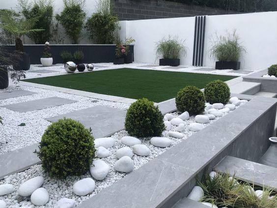 Nowoczesny ogród z geometrycznym trawnikiem, ścieżką z płyt i białych kamieni, kulistymi krzewami oraz minimalistyczną ścianą z bambusami w tle.