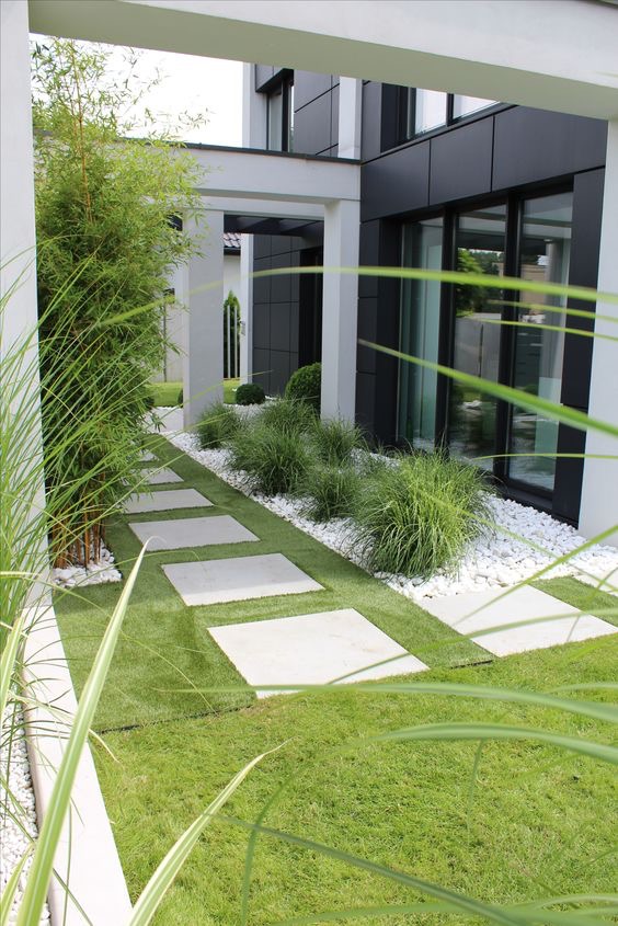 Nowoczesny ogród z minimalistyczną ścieżką z kwadratowych płyt betonowych na sztucznej trawie, obok budynku z ciemną elewacją i białymi ramami okiennymi, ozdobiony białym kamieniem i roślinami...
