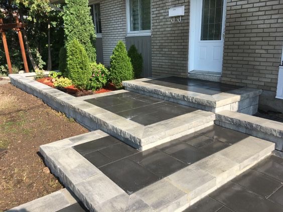 Schody wejściowe wyłożone szarą kostką brukową prowadzące do drzwi wejściowych domu z cegły, obok rabata z roślinami oddzielona niskim murem z kostki.