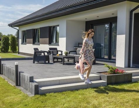 Nowoczesny taras z szarej kostki brukowej, schody z jasnoszarego kamienia, niskie ogrodzenie z betonowych elementów, czarne meble ogrodowe, młoda kobieta schodząca po schodach w letniej sukience