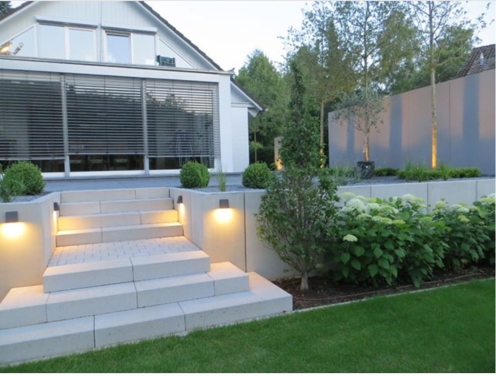 Eleganckie schody z jasnej kostki prowadzące do domu z szarymi żaluzjami, oświetlone lampami ściennymi, otoczone zielenią, w tym hortensjami i młodymi drzewami, z szarą ścianą w tle.