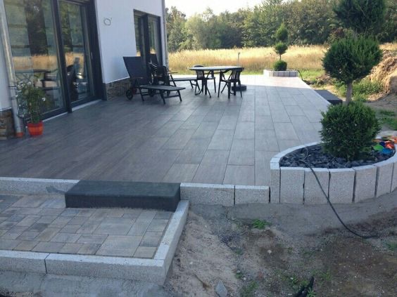 Nowoczesny taras wyłożony szarą kostką brukową, obrzeża z jasnego granitu, z elementami dekoracyjnymi w postaci kulistych krzewów i czarnych kamieni, widok na dom z dużymi oknami i ogród.