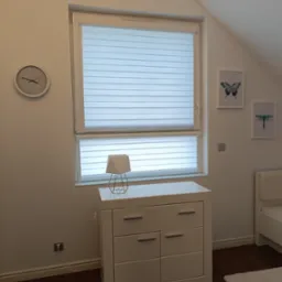 Pokój na poddaszu z oknem wyposażonym w poziome przesłony okienne, biała komoda z geometryczną lampką, zegar i grafiki na ścianie.