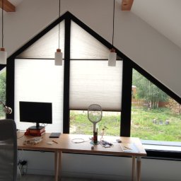 Sebastian Skoczylas - Poddasze z trójkątnym oknem osłoniętym plisowanymi zasłonami, biurko z komputerem i widokiem na zieleń za oknem.
