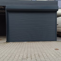 Sebastian Skoczylas - Zamknięta, grafitowa brama garażowa segmentowa z blachy falistej, widok z zewnątrz na tle jasnej elewacji budynku i kostki brukowej.