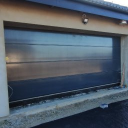 Sebastian Skoczylas - Czarna brama garażowa segmentowa, widok z zewnątrz, z widocznymi elementami montażowymi i przygotowaniem do wykończenia podjazdu.