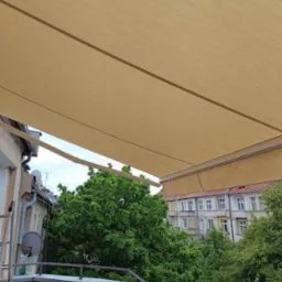 Beżowa, rozłożona osłona przeciwsłoneczna na balkonie z widokiem na zieleń drzew i budynki mieszkalne w tle, z drabina opartą o ścianę budynku.