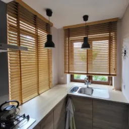 Jasna kuchnia z drewnianymi żaluzjami poziomymi w oknach, czarnymi lampami wiszącymi i srebrnym zlewem.