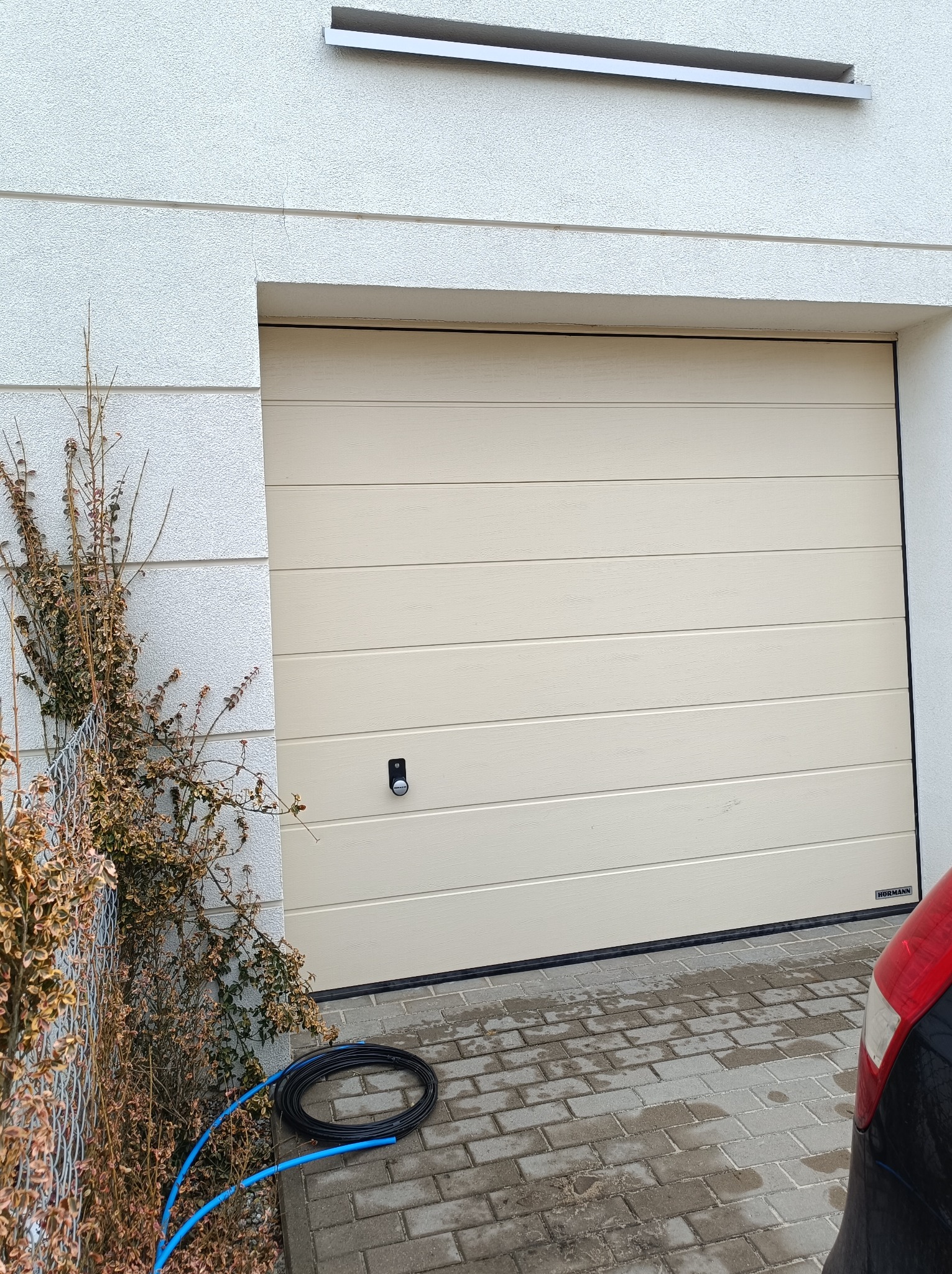 Beżowa segmentowa brama garażowa z automatycznym zamkiem, widoczna z zewnątrz, z fragmentem zaparkowanego samochodu i zwiniętym niebieskim wężem na kostce brukowej.