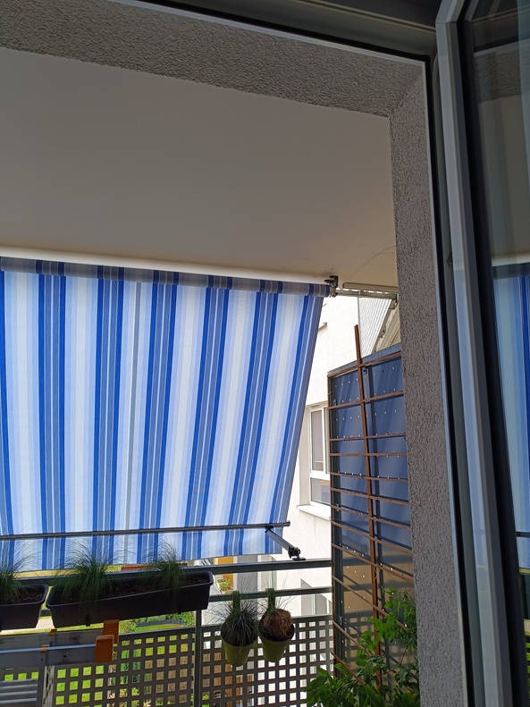 Balkon z częściowo rozwiniętą markizą w biało-niebieskie pasy, widok z wnętrza mieszkania, doniczki z roślinami na balustradzie.