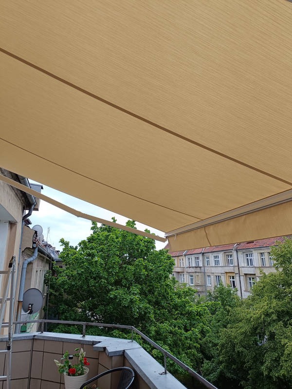 Beżowa, rozłożona osłona przeciwsłoneczna na balkonie z widokiem na zieleń drzew i budynki mieszkalne w tle, z drabina opartą o ścianę budynku.