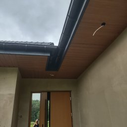 J.W.bud - Zamontowany system rynnowy z podsufitką z paneli drewnopodobnych, widoczny przewód elektryczny wystający z otworu w podsufitce, odbicie fotografa w szybie drzwi wejściowych.