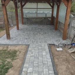 J.W.bud - Ułożona szara kostka brukowa pod drewnianą altaną z dekoracją w postaci czaszki jelenia, widoczne narzędzia brukarskie i niezakończone prace ziemne.