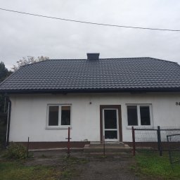 J.W.bud - Dom jednorodzinny z nowym, ciemnoszarym dachem z blachodachówki, widoczne rynny, komin i numer domu '84', na pierwszym planie fragment ogrodzenia i stos płyt.