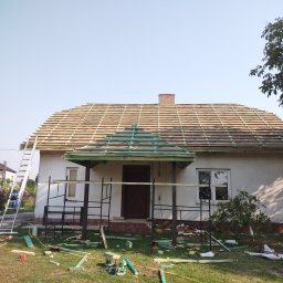 J.W.bud - Dom jednorodzinny z częściowo ułożoną konstrukcją dachu, widoczna więźba i łaty, rusztowanie przed wejściem, drabina oparta o ścianę, na trawie porozrzucane fragmenty drewna.