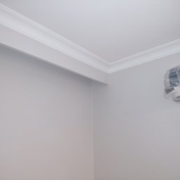 J.W.bud - Szare ściany z białą listwą dekoracyjną pod sufitem, klimatyzator w folii ochronnej w rogu pokoju.