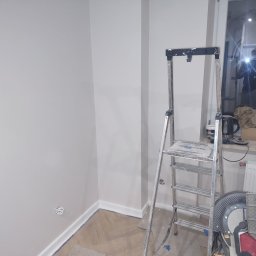 J.W.bud - Świeżo pomalowane, jasne ściany w rogu pokoju, widoczne listwy przypodłogowe, drabina malarska i narzędzia pracy, odbicie osoby robiącej zdjęcie w szybie okna.