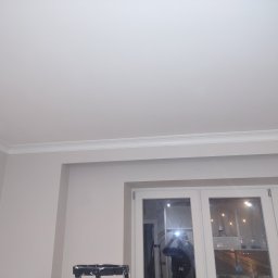 J.W.bud - Pomieszczenie po odświeżeniu: biały sufit z listwą dekoracyjną, ściany w odcieniu beżu, białe okno z widokiem na nocne miasto, drabina malarska w rogu.