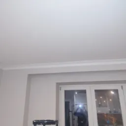 Pomieszczenie po odświeżeniu: biały sufit z listwą dekoracyjną, ściany w odcieniu beżu, białe okno z widokiem na nocne miasto, drabina malarska w rogu.