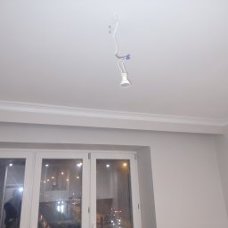J.W.bud - Biały sufit z listwą ozdobną i wiszącą żarówką na przewodach, widok na okno z odbiciem