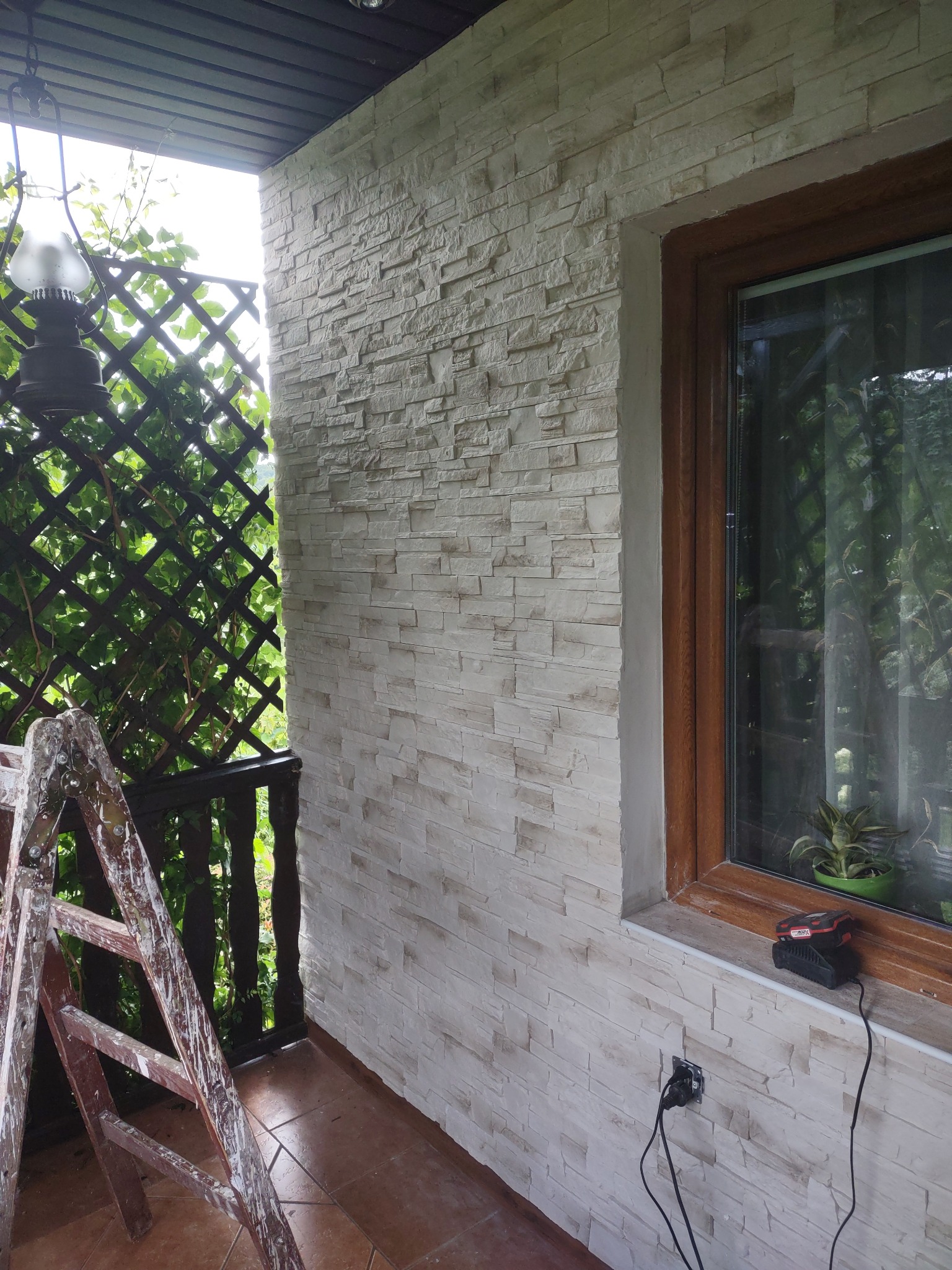 Ściana zewnętrzna wykończona jasnym kamieniem dekoracyjnym, z widocznym oknem w drewnianej ramie, drabina malarska i ładowarka akumulatorowa na parapecie.