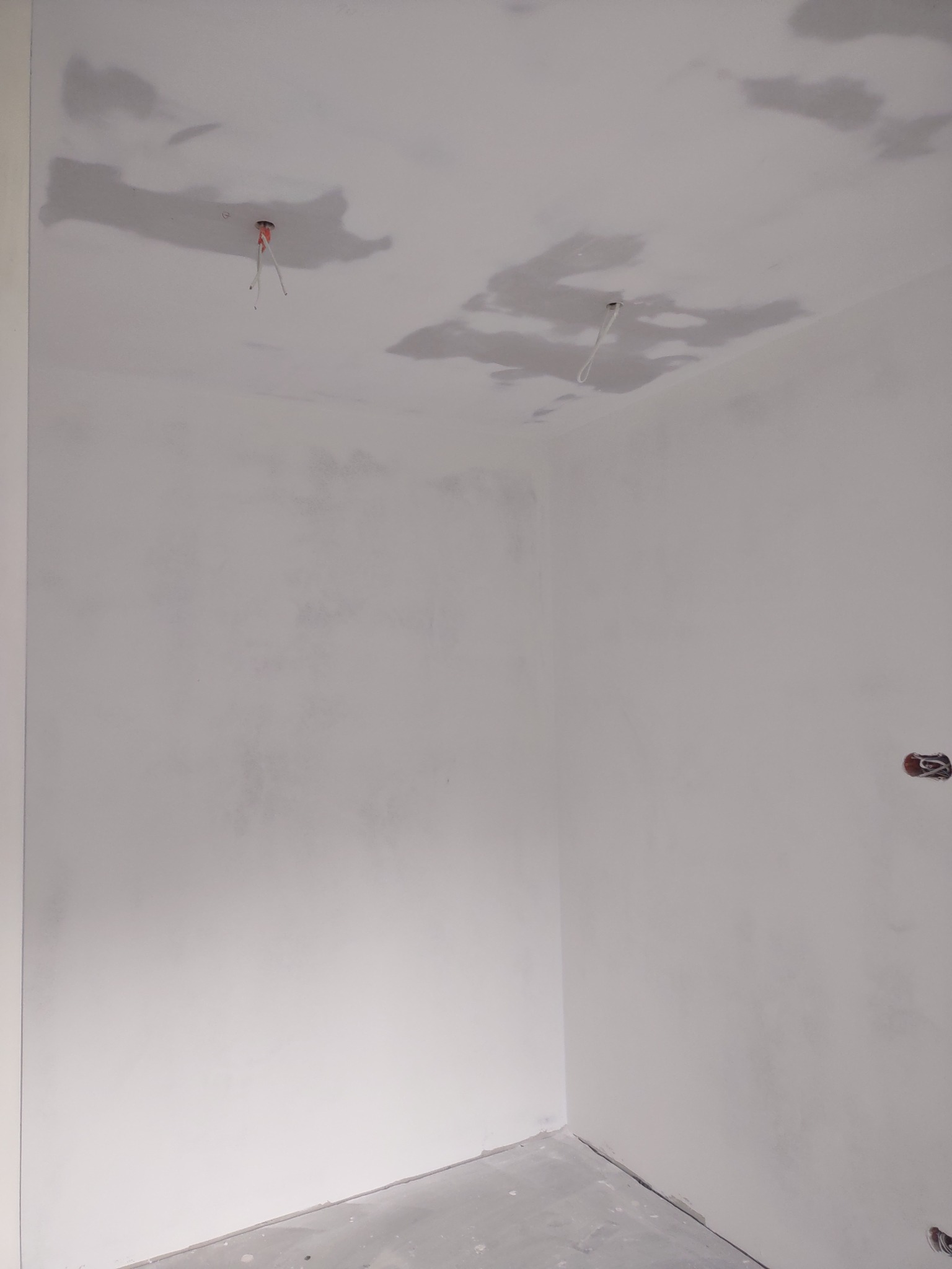 Pokój w trakcie prac wykończeniowych, widoczne ślady po szpachlowaniu na suficie i ścianach, wystające przewody elektryczne z sufitu, szara wylewka na podłodze.