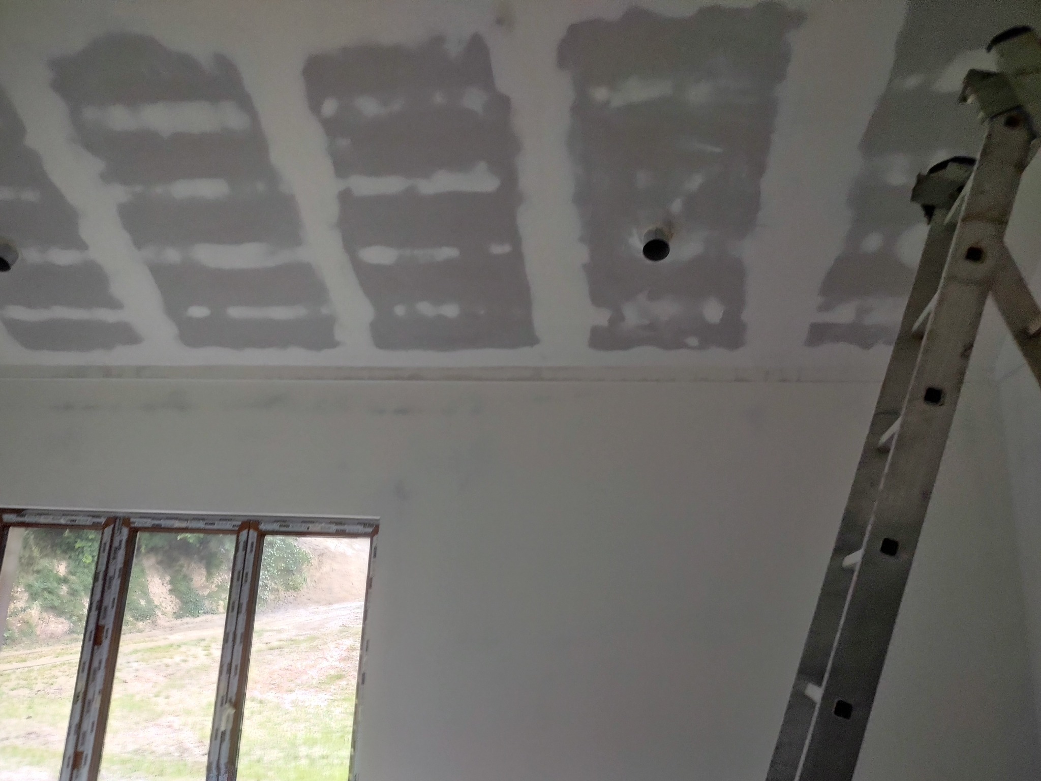 Wnętrze w trakcie wykańczania: sufit z widocznymi łączeniami płyt gipsowo-kartonowych przed szpachlowaniem, okno z widokiem na zieleń i drabina aluminiowa oparta o ścianę.