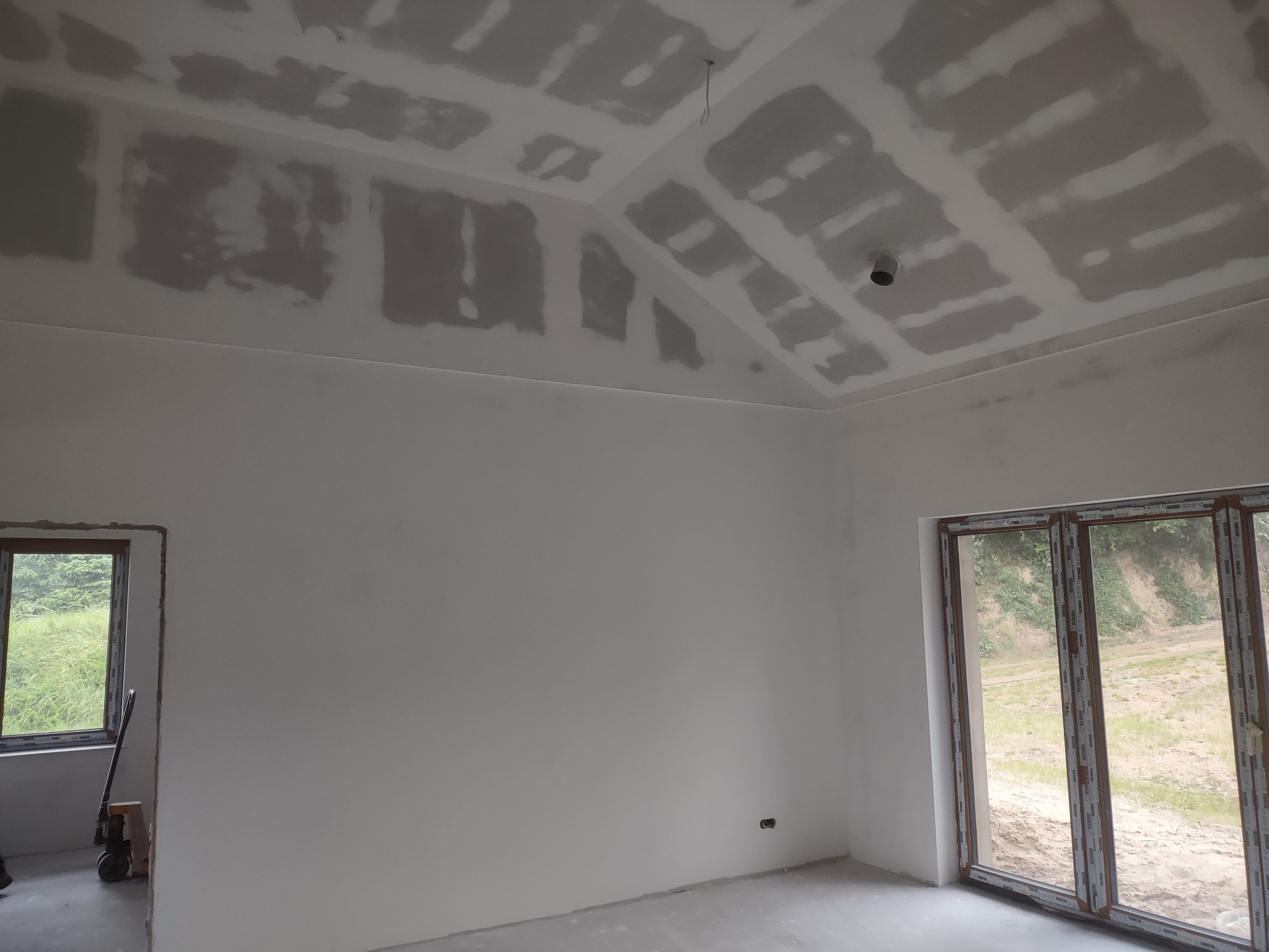 Wnętrze domu w trakcie prac wykończeniowych: ściany przygotowane do malowania, sufit z widocznymi łączeniami płyt gipsowo-kartonowych, okno z widokiem na zieleń na zewnątrz.
