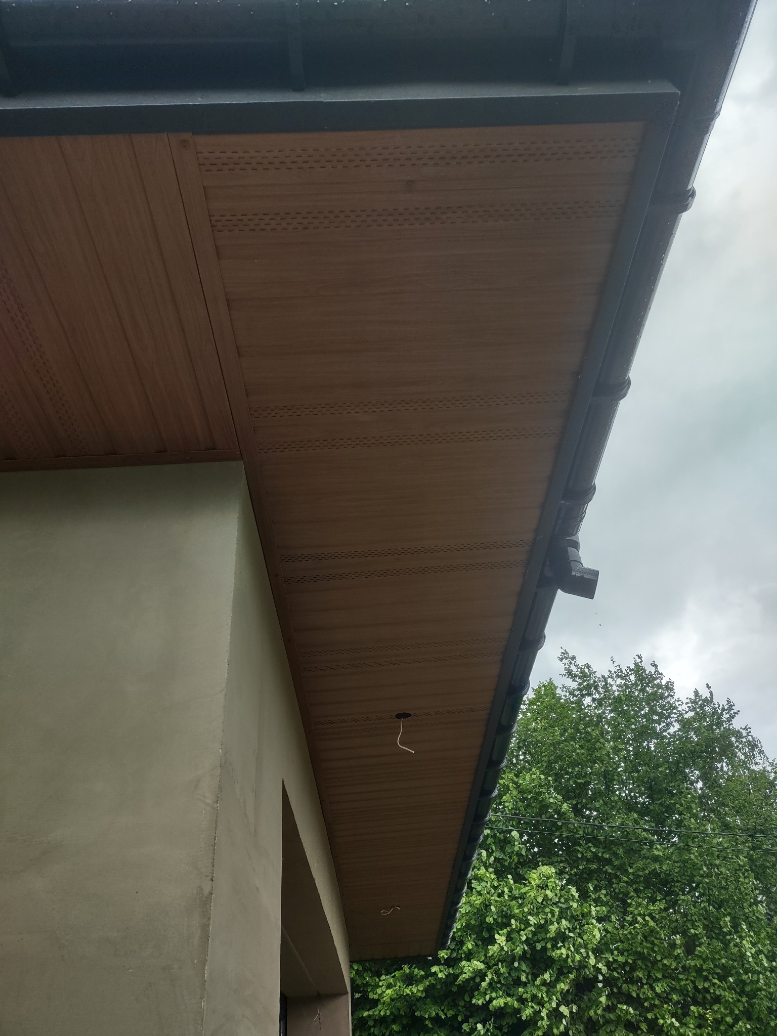 Drewnopodobna podbitka dachowa z widocznymi perforacjami i rynną, świeżo otynkowana ściana budynku w tle, widoczny fragment zieleni.