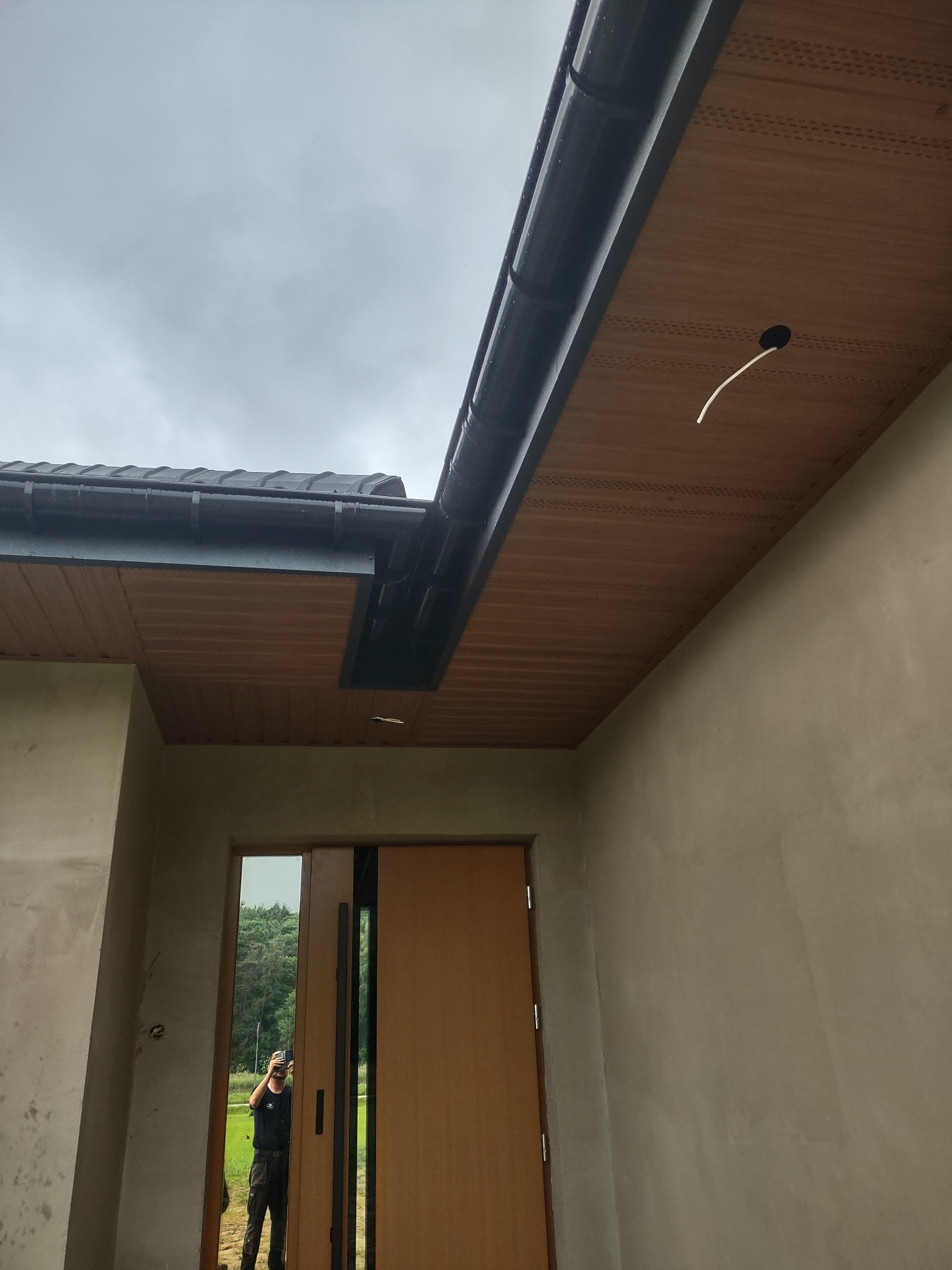 Zamontowany system rynnowy z podsufitką z paneli drewnopodobnych, widoczny przewód elektryczny wystający z otworu w podsufitce, odbicie fotografa w szybie drzwi wejściowych.