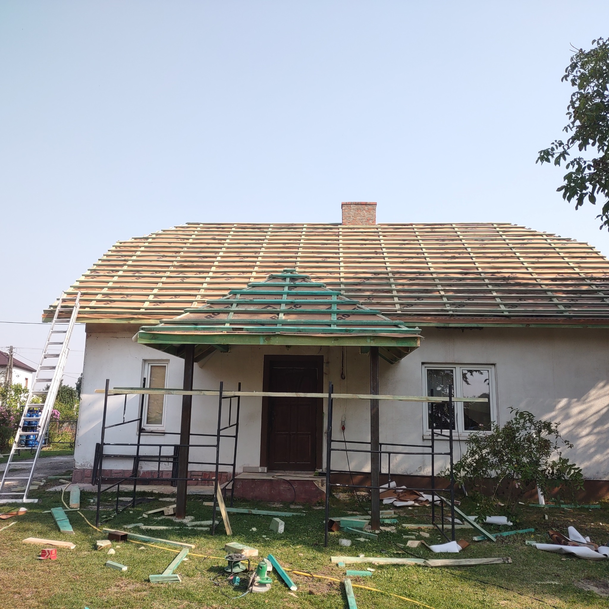 Dom jednorodzinny z częściowo ułożoną konstrukcją dachu, widoczna więźba i łaty, rusztowanie przed wejściem, drabina oparta o ścianę, na trawie porozrzucane fragmenty drewna.