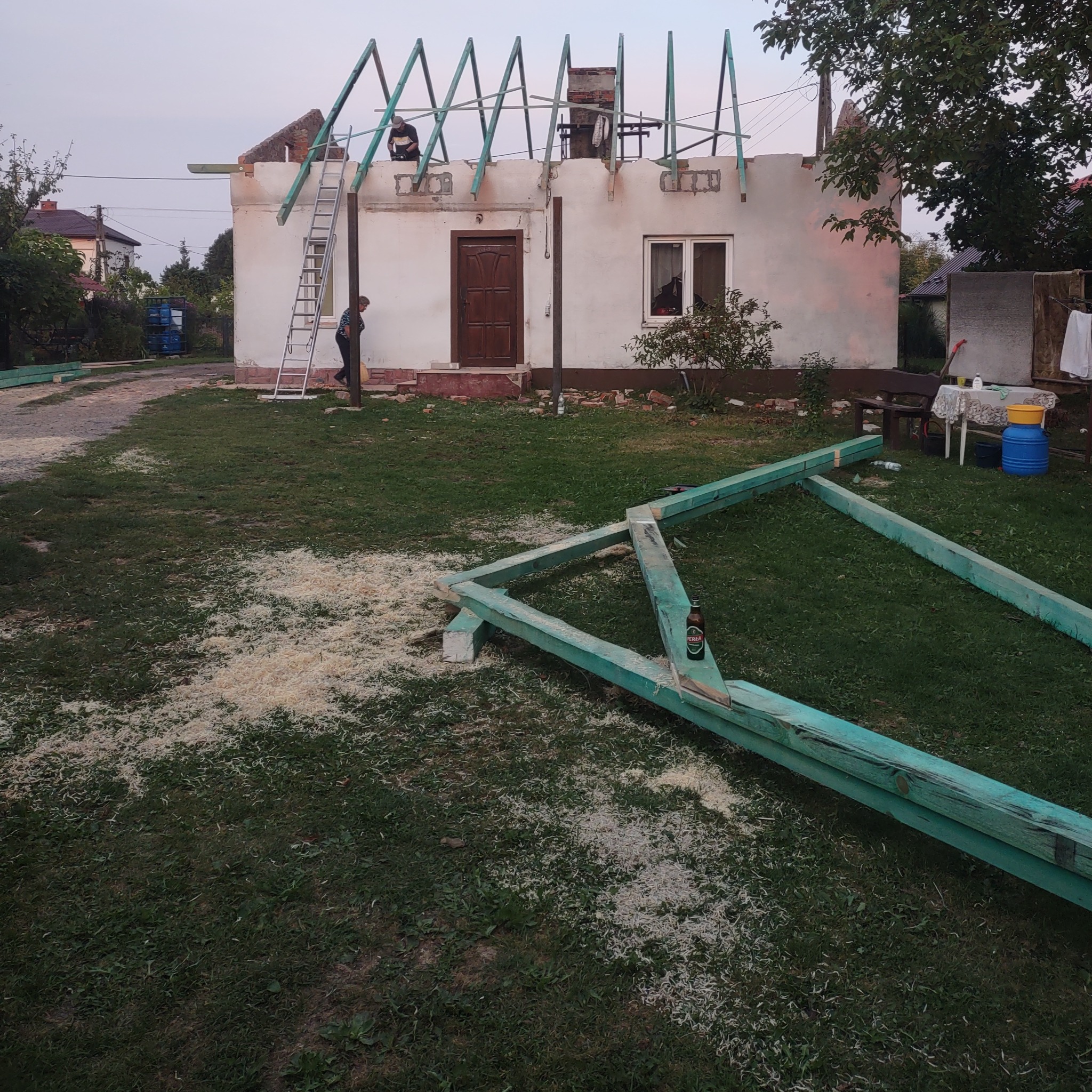 Dom w trakcie budowy, widoczna konstrukcja dachu z drewnianych belek, pracownicy na dachu, rozsypane trociny, butelka piwa Perła na belce, drabina oparta o ścianę budynku.