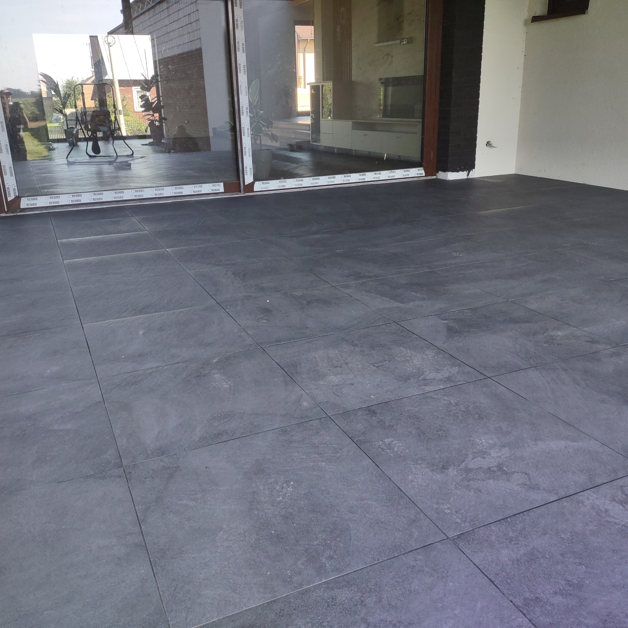 Wykończony taras z szarych, kwadratowych płytek ceramicznych, widok na przesuwne drzwi tarasowe z widocznym odbiciem ogrodu i wnętrza domu.