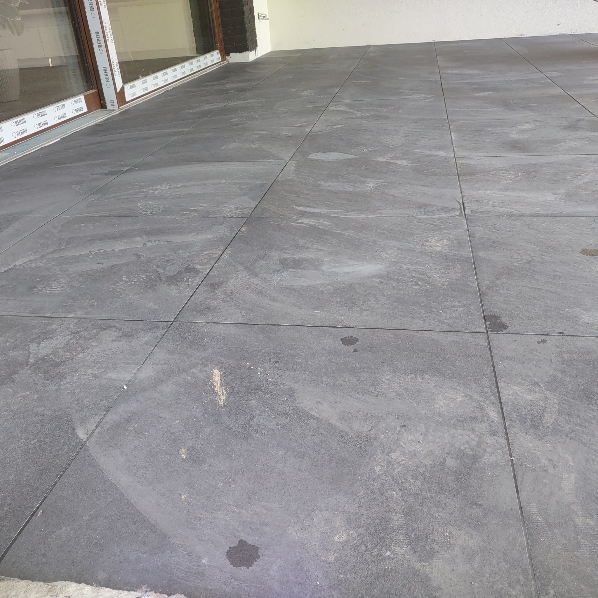 Szare płytki podłogowe ułożone na zewnątrz budynku, widoczne ślady zabrudzeń i niewielkie plamy, okno z brązową ramą w tle.