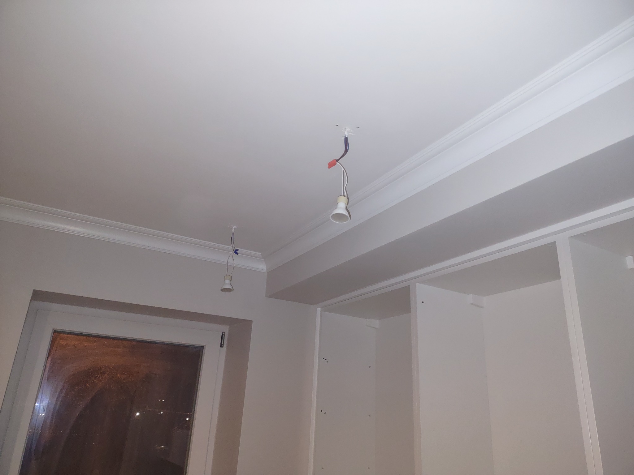 Wnętrze pokoju w trakcie prac wykończeniowych: biały sufit z listwami dekoracyjnymi, wiszące żarówki bez opraw, widoczne okno i fragment szafy w zabudowie.