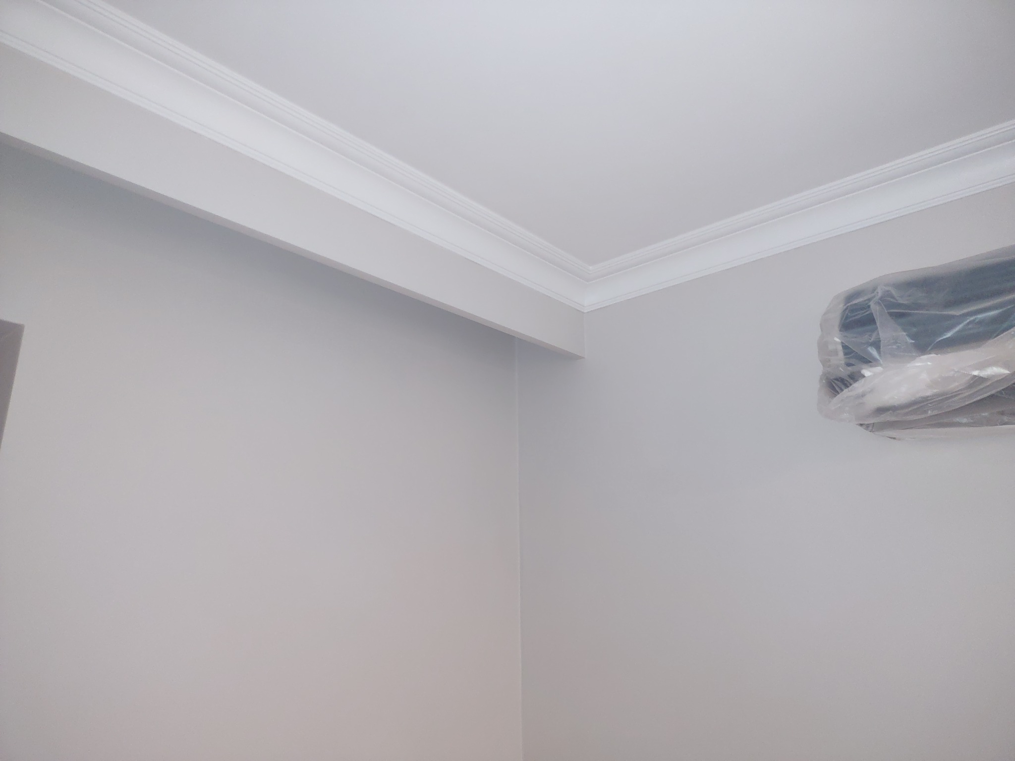 Szare ściany z białą listwą dekoracyjną pod sufitem, klimatyzator w folii ochronnej w rogu pokoju.