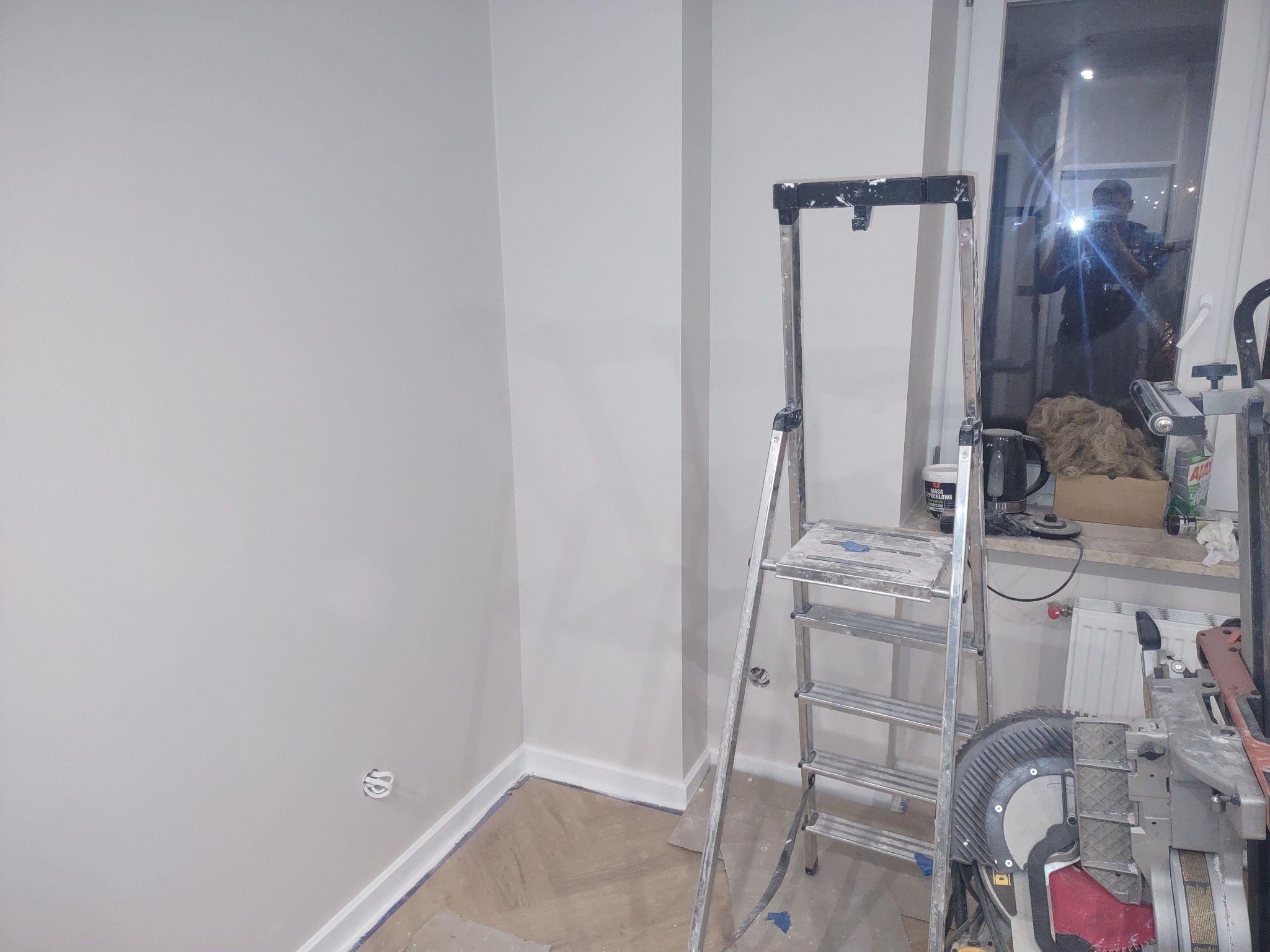 Świeżo pomalowane, jasne ściany w rogu pokoju, widoczne listwy przypodłogowe, drabina malarska i narzędzia pracy, odbicie osoby robiącej zdjęcie w szybie okna.