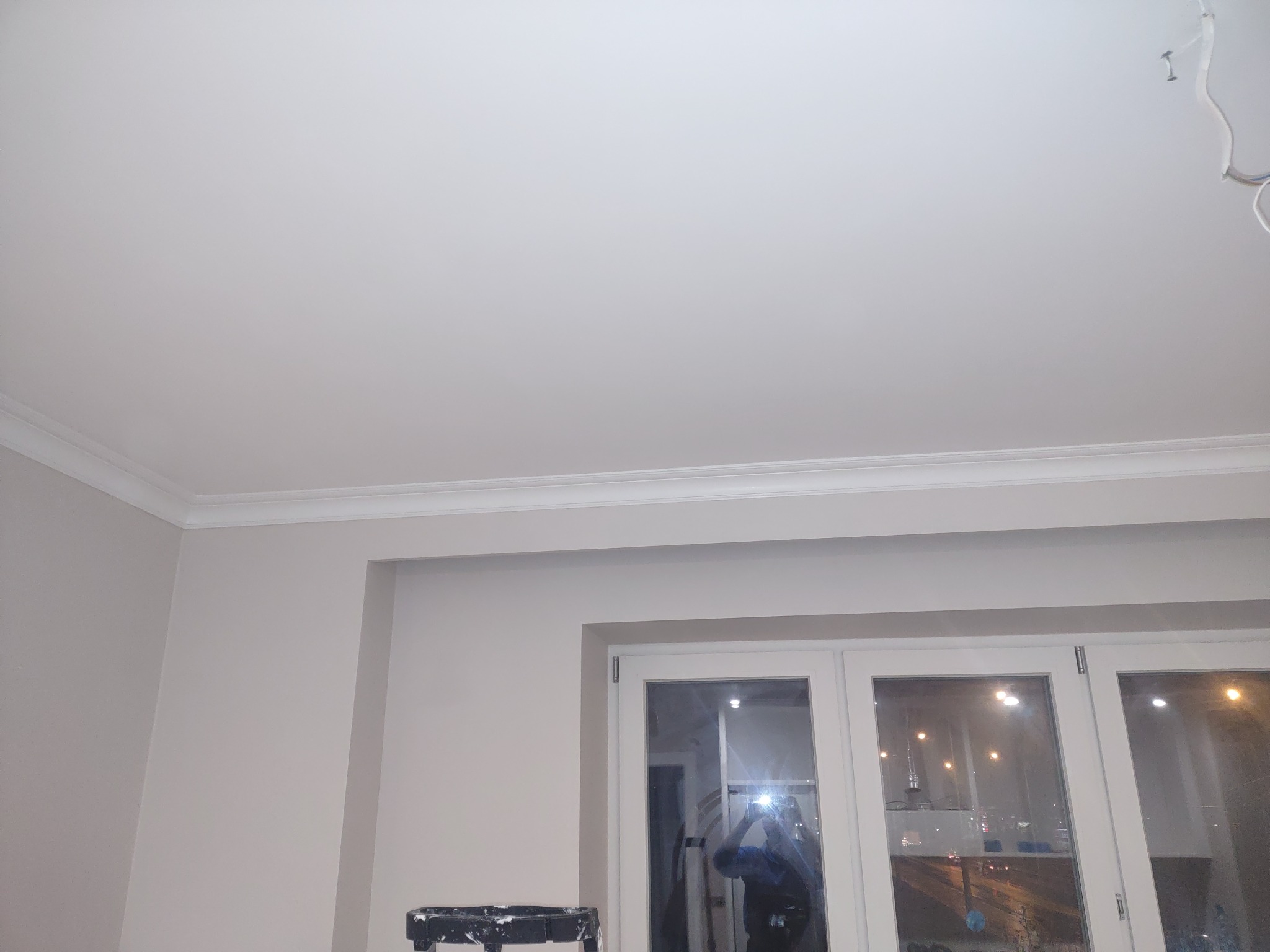 Pomieszczenie po odświeżeniu: biały sufit z listwą dekoracyjną, ściany w odcieniu beżu, białe okno z widokiem na nocne miasto, drabina malarska w rogu.