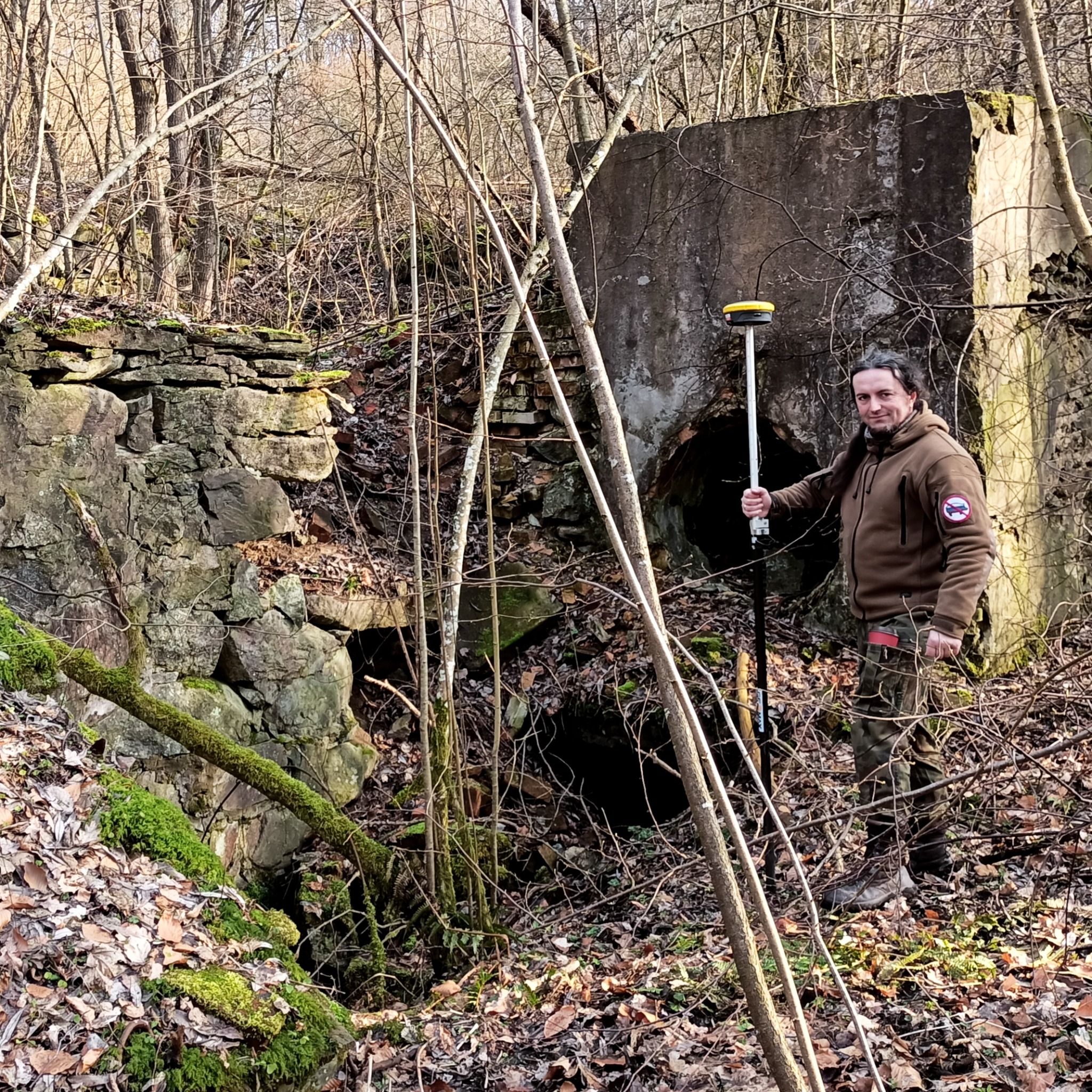 Mężczyzna z odbiornikiem GNSS w terenie z ruinami, pomiary geodezyjne w zaroślach, widoczne stare mury z kamienia i betonowy bunkier.