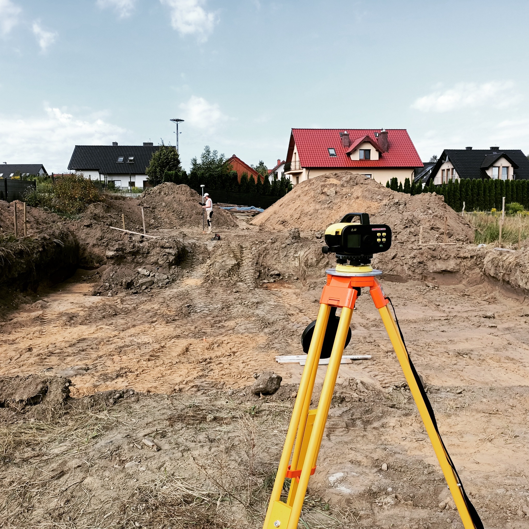 Żółty niwelator ustawiony na placu budowy domu jednorodzinnego, w tle widoczny pracownik w białym kasku i wykop fundamentowy.