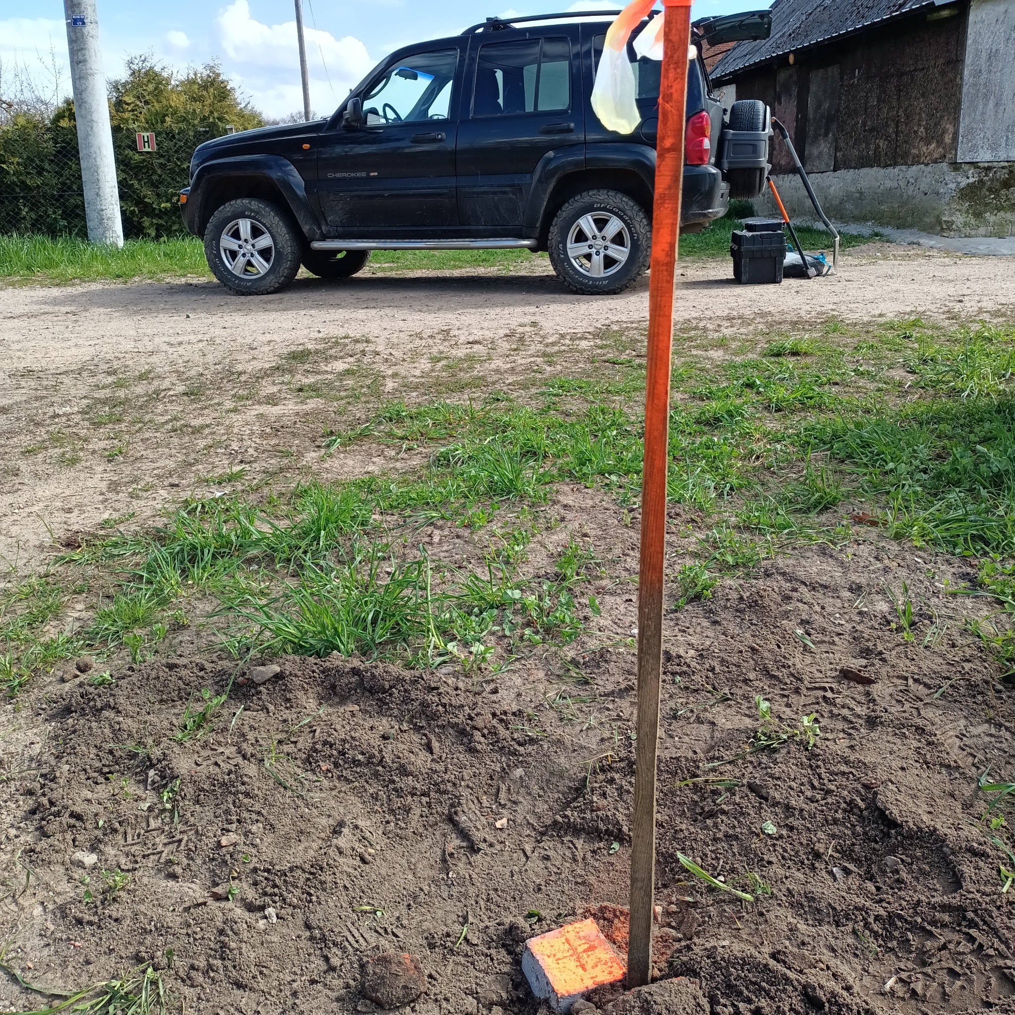 Wbity w ziemię pomarańczowy palik z zawieszoną białą folią, obok czarny samochód terenowy i narzędzia geodezyjne na tle wiejskiego krajobrazu.