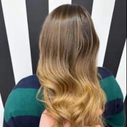 Włosy ombre z brązowych przechodzące w blond, delikatne fale, widok z tyłu, na tle ściany w czarno-białe pasy i ręka trzymająca pasmo włosów.