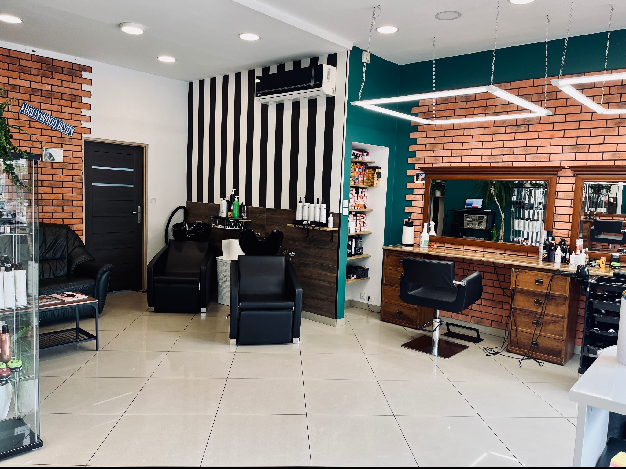 Wnętrze salonu z dwoma stanowiskami do mycia włosów, czarnymi fotelami i ścianą z cegły oraz napisem Hollywood Blvd.