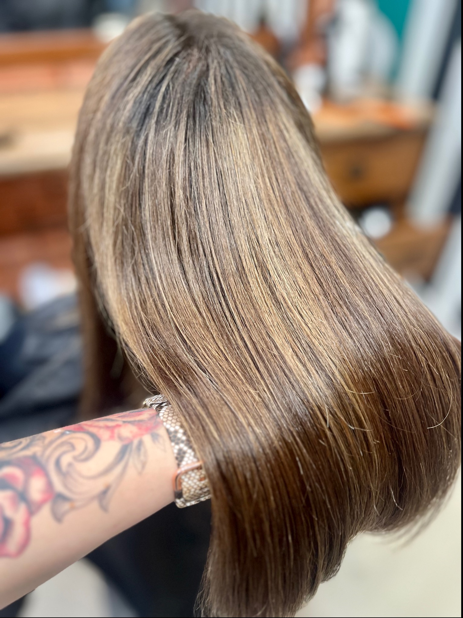 Proste, lśniące włosy w odcieniach brązu i blondu, widok z tyłu, ramię z tatuażem i bransoletką w kadrze.