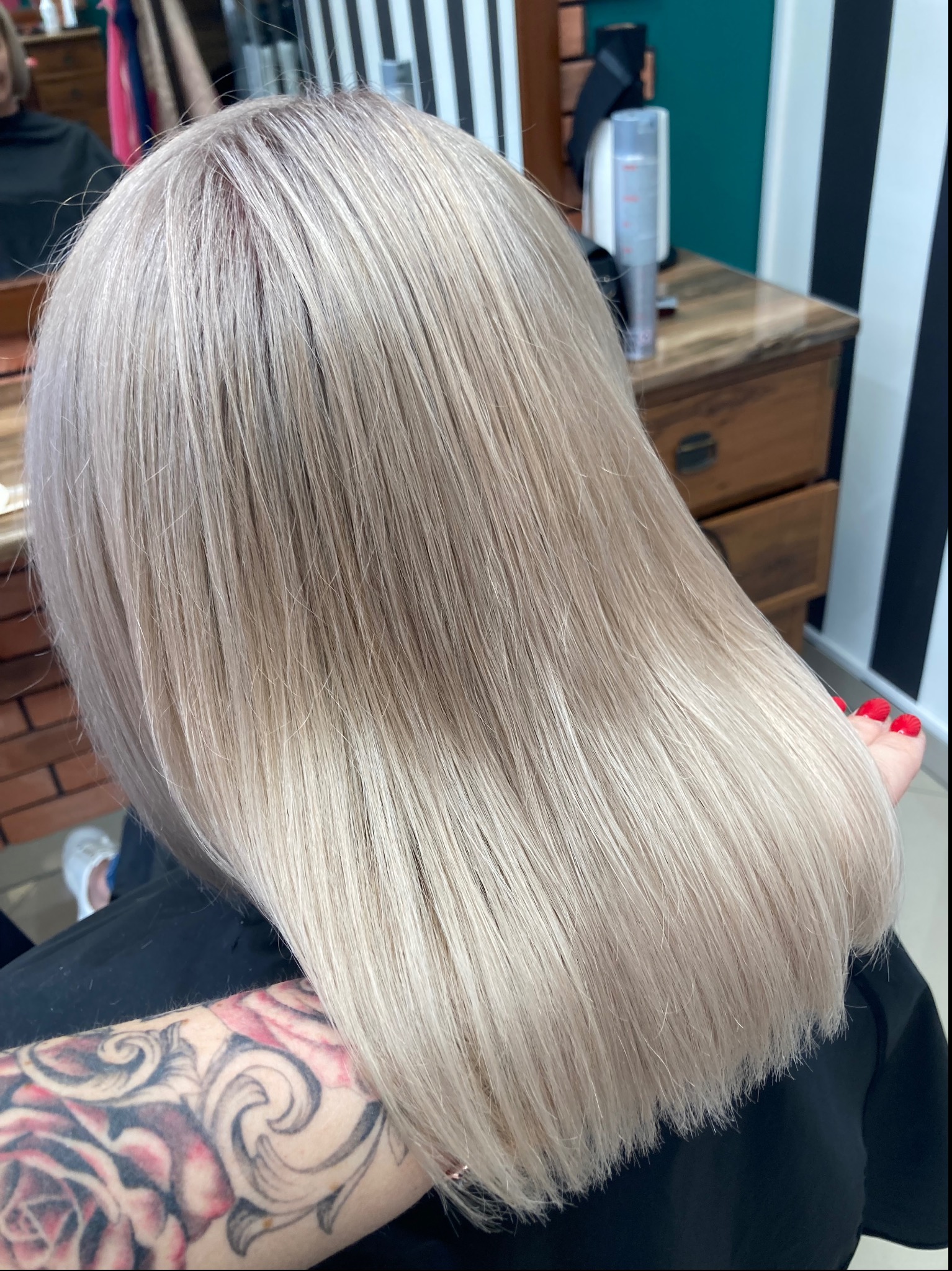 Świeżo ostrzyżone, proste, platynowe blond włosy z ciemniejszym odrostem, widoczne tatuaże na ramieniu klienta, w tle elementy wyposażenia salonu.