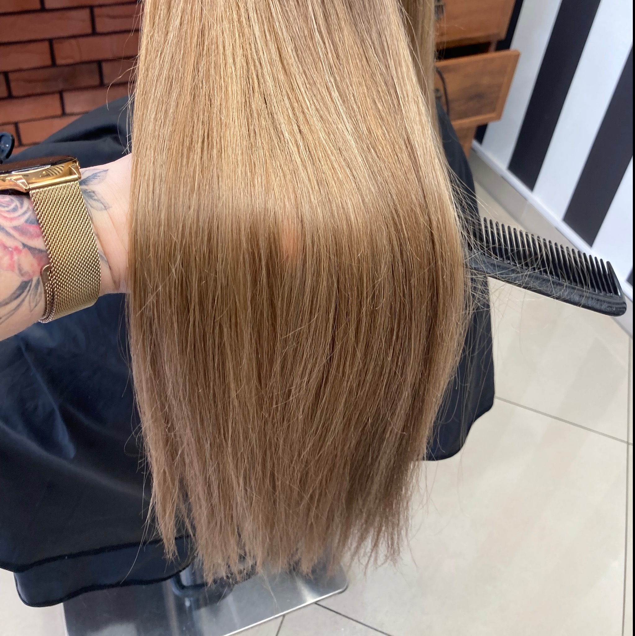 Proste, blond włosy rozczesywane czarnym grzebieniem, widoczna ręka z tatuażem i złotym zegarkiem na tle ceglanej ściany i czarno-białych pasów.